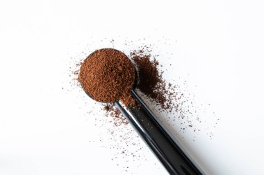 Beyaz arka planda bir kaşık içinde öğütülmüş kahve, etrafına dağılmış kahve tozu.