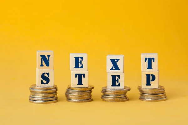 Next Steps concept with wooden block on step stacked coins on yellow background.