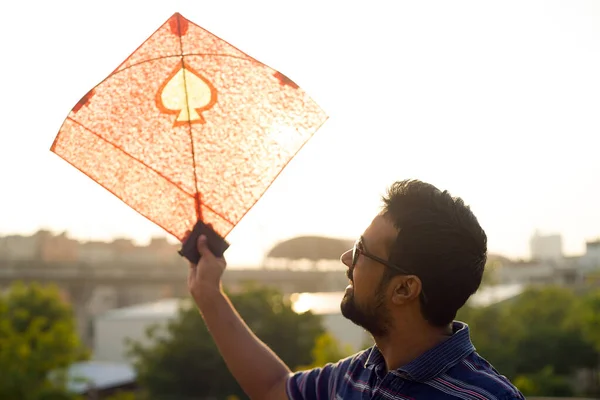 Young man holding aloft colorful paper and wood kite against a blurred background setting sun on the indian kite festival of makar sankranti or uttarayana — Stock Photo, Image