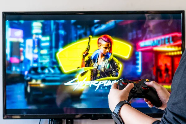 Mulher segurando um controlador de engrenagem vermelha e jogando popular jogo de vídeo Cyberpunk 2077 em uma televisão e PC — Fotografia de Stock