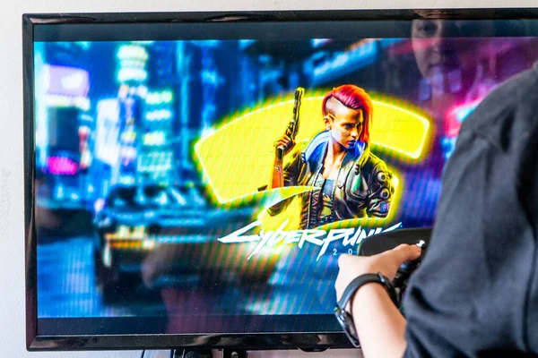 Mujer sosteniendo un controlador de vapor y jugando popular videojuego Cyberpunk 2077 en una televisión y PC — Foto de Stock