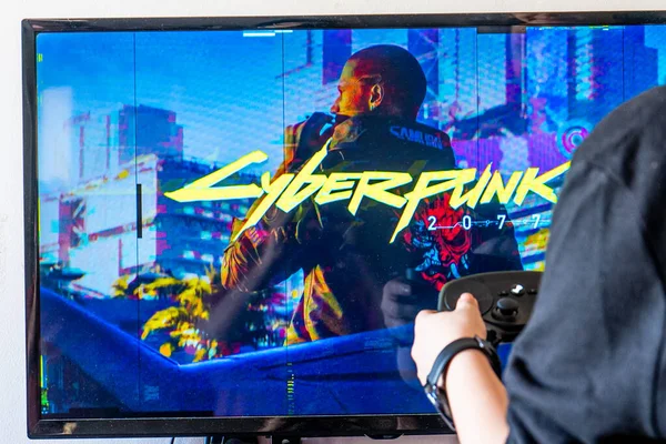 Mujer sosteniendo un controlador de vapor y jugando popular videojuego Cyberpunk 2077 en una televisión y PC — Foto de Stock