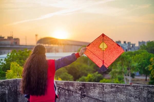 Młoda kobieta trzyma wysoko kolorowy papier i drewno latawiec przed zamazanym tle zachodzące słońce na indyjskim festiwalu latawca makar sankranti lub uttarayana — Zdjęcie stockowe