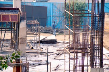 Kapüşonlu ve miğferli insan işçilerinin bulunduğu Hint şantiyesi gökdelen inşaatı için yarım kalmış kiriş sütunlarının yanında ekipman taşıyor.