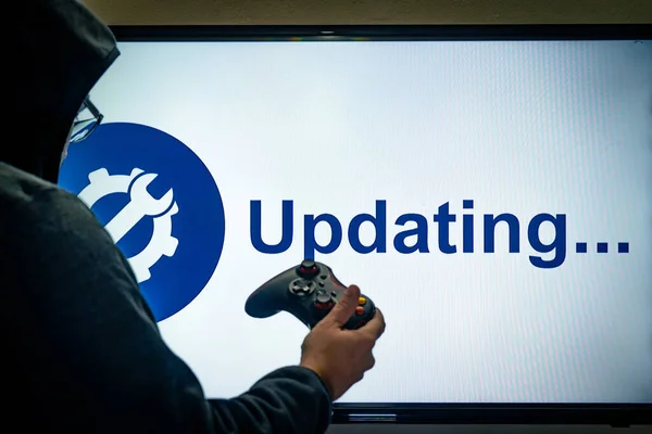 Hombre encapuchado sosteniendo un controlador de juego delante de una pantalla que muestra un mensaje de actualización para un juego o software día 1 parche —  Fotos de Stock