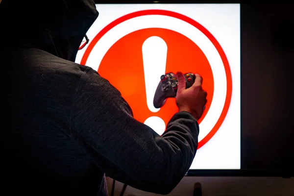 Man in hood holding controller in front of screen showing an alert red exclamation message as a game crashes due to bugs, hacks or issues — Stock Photo, Image
