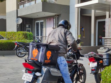 Motosikletli, siyah ve turuncu şeritli yiyecek çantalı tek boynuzlu at firması için yiyecek ve sebze teslimatı yapacak.