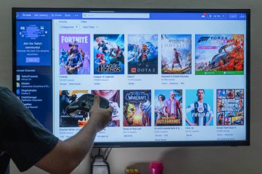 PC konsol ekranının önünde buhar kumandası tutan adam, insanların para kazanmak için tam zamanlı yayın yaptığı oyunlarla birlikte seğirme platformunun ana ekranını gösteriyor.