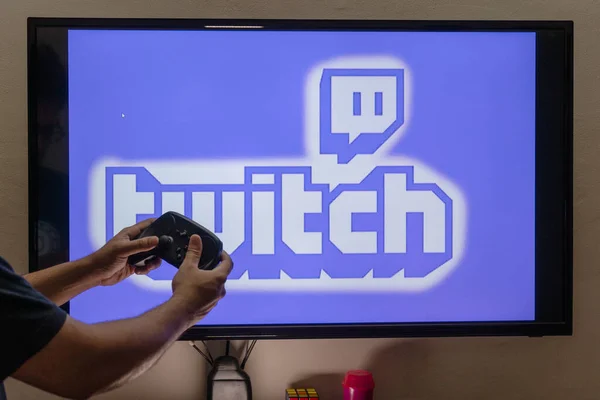 Hombre sosteniendo un control remoto de vapor frente a una pantalla de consola de PC que muestra la plataforma de transmisión de contracción donde la gente está transmitiendo a tiempo completo para ganar dinero —  Fotos de Stock