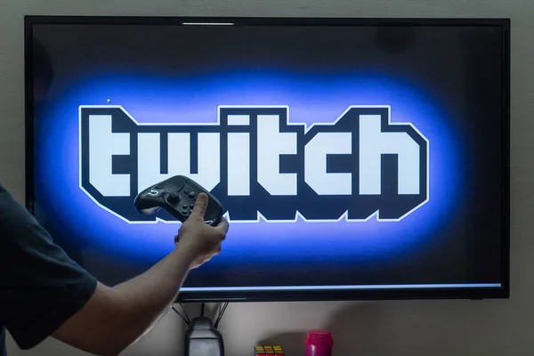 Hombre sosteniendo un control remoto de vapor frente a una pantalla de consola de PC que muestra la plataforma de transmisión de contracción donde la gente está transmitiendo a tiempo completo para ganar dinero —  Fotos de Stock