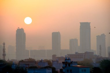 Etrafı sisli gökdelenlerin alacakaranlık görüntüsü ve arkalarında güneş doğarken Gurgaon Delhi 'de Hindistan' da ön planda küçük evler var.