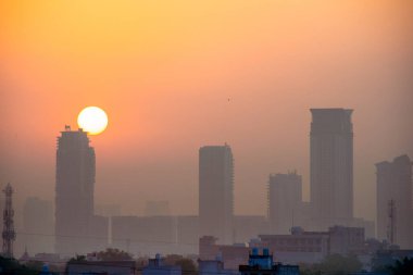 Etrafı sisli gökdelenlerin alacakaranlık görüntüsü ve arkalarında güneş doğarken Gurgaon Delhi 'de Hindistan' da ön planda küçük evler var.