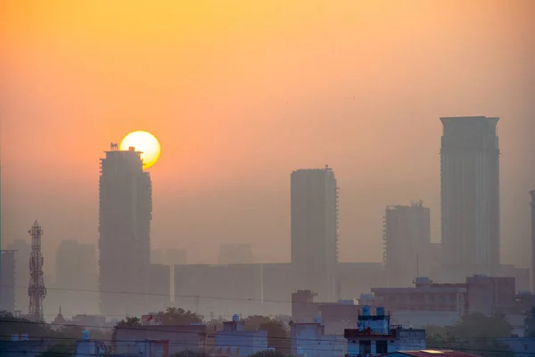 在印度的古尔冈，黄昏的黎明时分，摩天大楼周围都是雾，太阳升起在他们身后，前景是一些小房子 — 图库照片