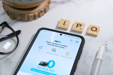 Yakın bir yere yerleştirilmiş kalem ve gözlüklerle cep telefonunda paytm logo ekranı nakitsiz ödemeler için tek boynuzlu at ödeme hizmetleri sağlayıcısının halka açılışını gösteriyor