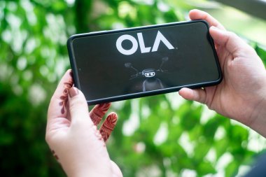 Cep telefonunda Ola Electric 'in Asya' daki yeni S1 elektrikli scooter uygulamasını gösteren bir cep telefonu var. Dışarıda bitkilere karşı telefonu tutan genç bir kadınla.
