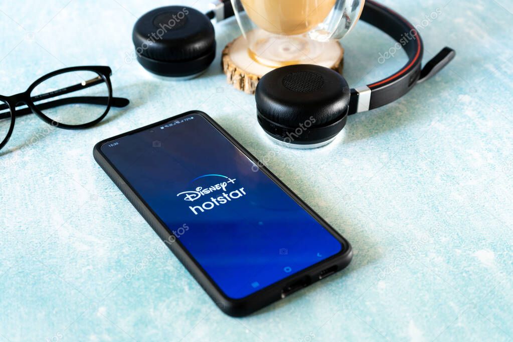 Mumbai, India - circa 2021: Mobile phone smartphone with popular OTT streaming app hotstar Disney plus in India. Flatlay shot with headphones specs and a cup of coffee