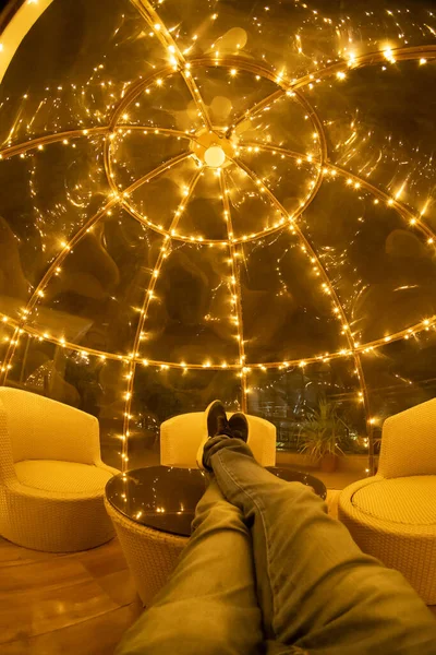 Indoor fisheye tiro de homem sentado em uma cúpula de vidro de plástico com luzes ao redor com sofás brancos e pernas para cima na mesa de vidro relaxante em um retiro natural — Fotografia de Stock