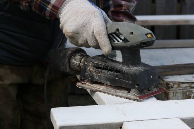 Elektrik titreşimli zımpara makinesi olan bir adam bir tahtanın yüzeyini tedavi eder.