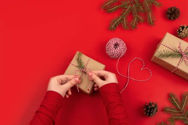 Kız elleri Noel için hediye kurdelesine fiyonk bağlar. El işi kâğıdı ve parşömende paketlenmiş hediye, kırmızı arka planda ladin dalları.