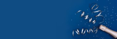 Photo banner of champagne bottle with confetti stars and party streamers on trendy classic blue background. Christmas, birthday or wedding concept. Flat lay, top view, copy space, mock-up.