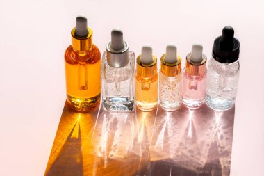 Farklı cam şişelerde güzellik serumu ya da yağ ya da güneş ışığında pastel pembe arka planda gölgeli losyon ürünleri. Güzellik kavramı, kozmetoloji, dermatoloji. Ön görünüm, boşluğu kopyala.