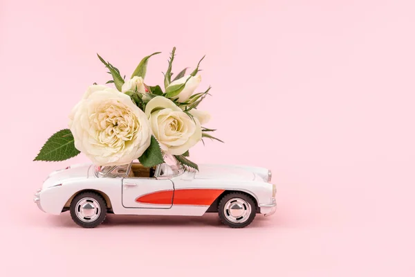 Retro white car delivering bouquet of white rose flowers on pink background. February 14, Valentine\'s day, 8 March, International Women\'s Day. Flower delivery. Copy space.