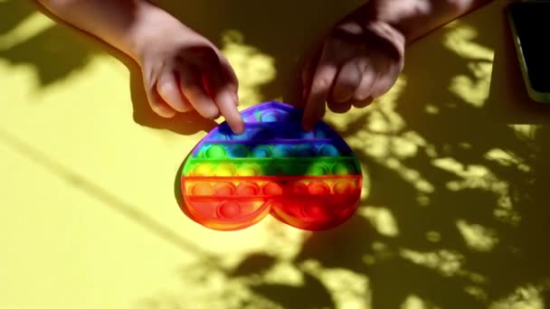 Vista superior de las manos de un niño jugando con un Popit en color arco iris y forma de corazón sobre fondo amarillo. — Vídeo de stock