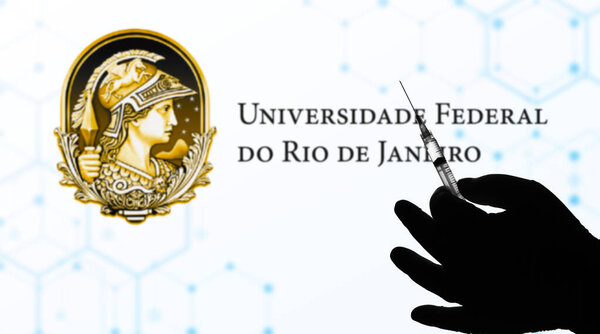 August 11, 2021, Brazil. In this photo illustration a hand holding a medical syringe is seen with Universidade Federal do Rio de Janeiro (UFRJ) company logo displayed on a screen in the background