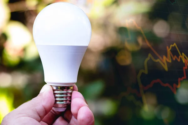 Hand holding a LED light bulb (light-emitting diodes). In the background, the graph of the stock exchange. Electricity price concept, readjustment in the electricity bill