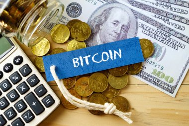 Bitcoin kelimeleri, yüz dolarlık banknotlar, altın sikkeler ve ahşap arka plan üzerinde hesap makinesi.