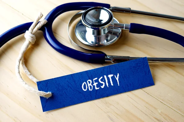 Fettleibigkeit Wort auf Etikett und Stethoskop auf Holzgrund geschrieben. — Stockfoto