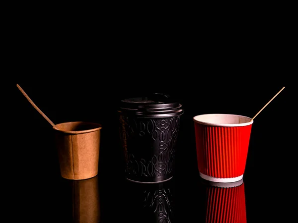 Disposable cardboard coffee cups on a black background. Cardboard colored cups. Disposable tableware. Coffee drinks. Paper dishes for drinks. Hot tea. Black background. Darkness.