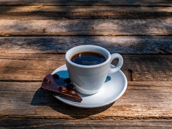 A cup of coffee and dark chocolate in a saucer on a wooden table. Coffee drink. Dark chocolate bar. Cup and saucer. Wooden table from a bar. Food and drinks. Ceramic tableware. Street Cafe.