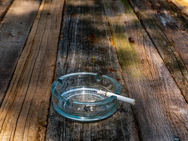 Glass ashtray with a cigarette on a wooden table. Smoking area. A cigarette in an ashtray. Wooden table. Tobacco products. Bad habit. Nicotine. Health and medicine. The smoker is passive.