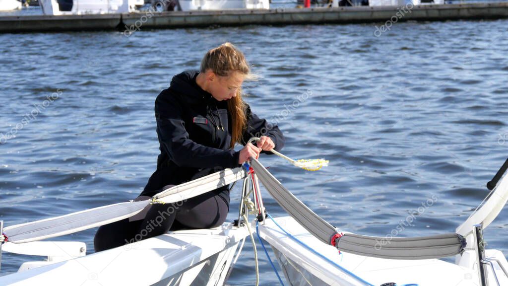 A young athlete, a blonde girl in a tracksuit, is mooring a yacht on the river on a cool, windy day.