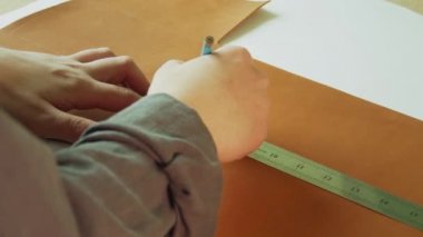 Yavaş çekimde bir terzinin elleri, bir kalem ve cetvelle doğal kahverengi deri parçasına yakın çekim işaretleme ve kesme çizgisi çiziyor..