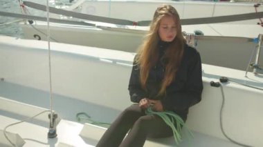 Beyaz bir spor yatının güvertesinde oturan uzun saçlı bir kız tekneyi çoklu gün yelkenli yarışına hazırlamak için ipi çözüyor..