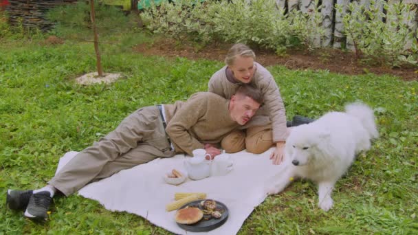 Молода пара чоловік і жінка на пікніку в саду зі своїм білим собакою в сповільненому темпі — стокове відео