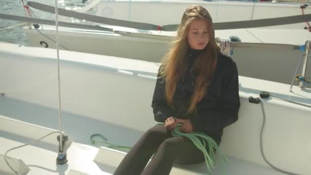 Дівчина з довгим волоссям, що сидить на палубі білої спортивної яхти, розмотує мотузку, щоб підготувати човен для участі в багатоденній регаті . — стокове відео