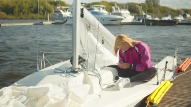 Μια νεαρή όμορφη αθλήτρια ιστιοπλοΐα ελέγχει την πλοήγηση στο smartphone της, ενώ κάθεται στο κατάστρωμα ενός γιοτ στην προβλήτα. — Αρχείο Βίντεο