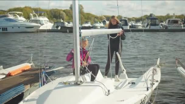 Deux filles dans des coupe-vent sportifs sur le quai attachent le gréement au boom d'un yacht blanc rapide. — Video