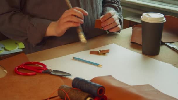 Un sastre experimentado en el taller detrás del banco de trabajo prepara accesorios para crear una bolsa de cuero. Une elementos y bebe café durante el descanso. — Vídeo de stock