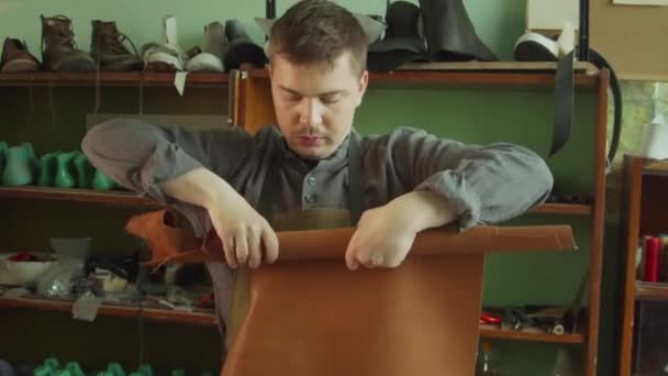 En skomakarlärling rullar en bit äkta läder i en rulle. I bakgrunden, rack med blanksteg för skor. — Stockvideo
