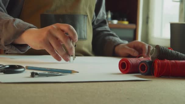 Un sastre en un delantal hace una marca con brújulas en el patrón. En primer plano, carretes con hilos de colores. — Vídeos de Stock