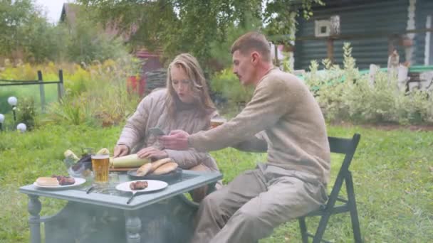 De man drinkt bier en laat zijn vrouw tijdens de lunch grappige foto 's zien op een smartphone in de dacha — Stockvideo