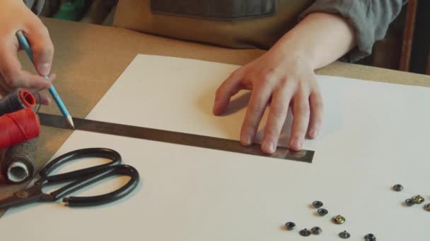 Vista dall'alto le mani di un maestro calzolaio fanno un disegno a matita degli elementi di una borsa di pelle su carta. Nella cornice di matasse di filo e forbici. — Video Stock