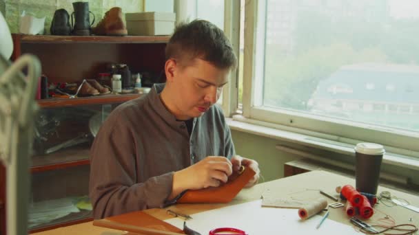 A young skilled tailor sews the element to a leather bag using a tailors needle. In the interior of a leather workshop with a window. — Stock Video