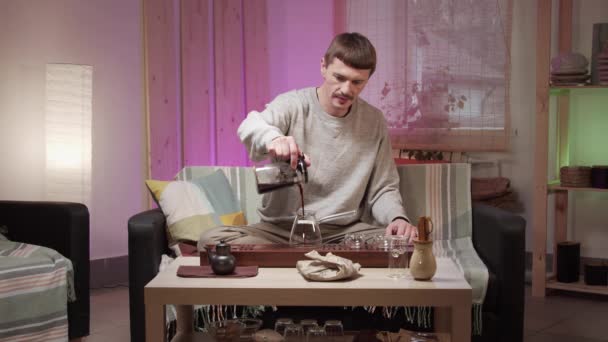 Чоловік середнього віку, що тримає чайник вареного чаю, випиває напій у скляну чашку і п'є чай — стокове відео