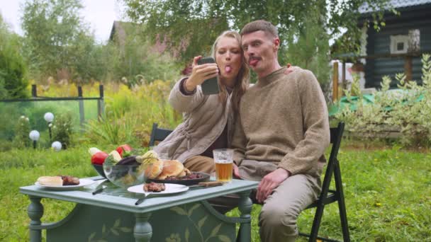 Een jong stel neemt een selfie aan een picknicktafel in het land op een smartphone, laat de taal zien. Langzame beweging — Stockvideo