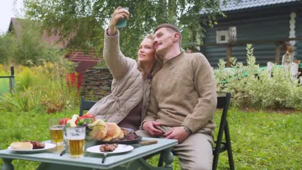 Een jong stel brengt de zomer in het land door in zelfisolatie, neemt een selfie op smartphone, de vrouw kust haar man — Stockvideo